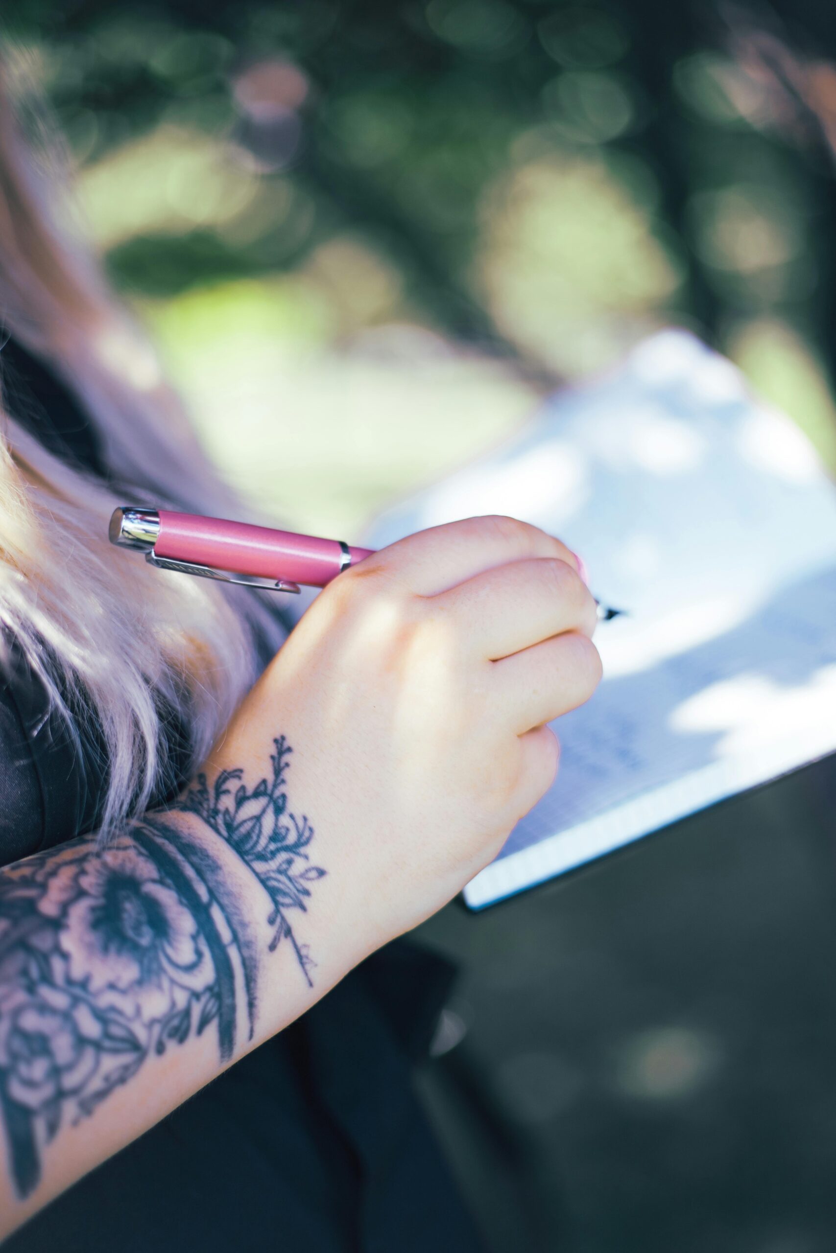 Close-up of a tattooed arm while a woman writes in a notebook outdoors.